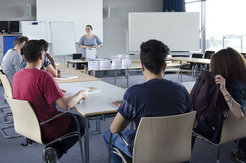 Participants sit in a seminar room while a person at the front of the table presents materials.